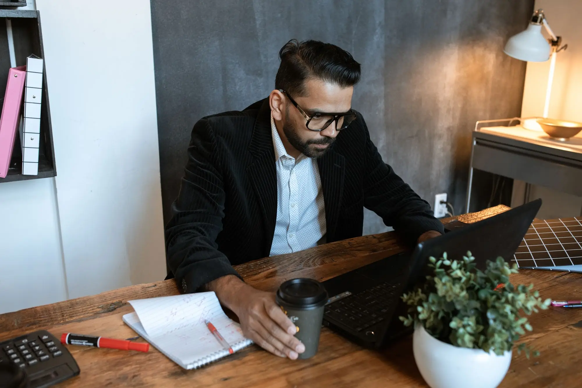 Company director in a dark jacket working on a laptop at a timber desk with a notebook and takeaway coffee, sorting out how to pay himself from the business