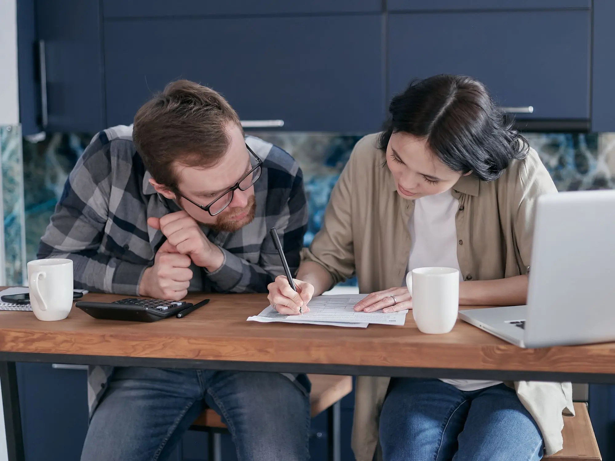 Couple at their kitchen bench working through home loan paperwork with a calculator and laptop beside them